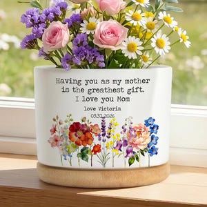 Puede incluir: Una maceta de cerámica blanca con base de madera, llena de un vibrante arreglo de rosas rosas, statice morado y margaritas blancas. La maceta tiene un diseño floral y el mensaje: "Having you as my mother is the greatest gift. I love you Mom."