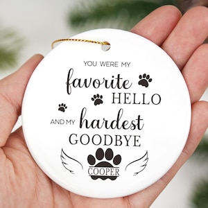 May include: A white ceramic disc ornament with a gold hanging loop. The text "You were my favourite HELLO and my hardest GOODBYE" is printed on the front, along with paw prints and angel wings. The name "COOPER" is below the wings.