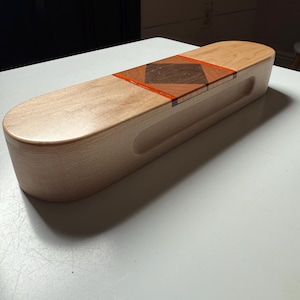 Puede incluir: Una caja de madera de color claro con una parte superior redondeada y un asa empotrada. La tapa presenta una incrustación decorativa de madera oscura y detalles en naranja. La caja es rectangular y parece hecha a mano.