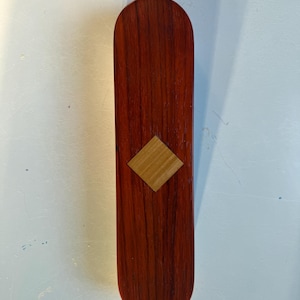 May include: A long, rectangular wooden box with a reddish-brown finish. A light-colored, diamond-shaped inlay is centered on the front. The box has rounded ends and appears to be made of a hardwood.
