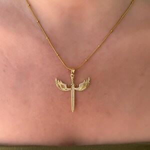 May include: A gold-colored necklace featuring a pendant with a sword design, adorned with small, sparkling stones. The sword is flanked by detailed wings, and the chain has small, evenly spaced beads. The necklace is displayed against a skin-toned background.