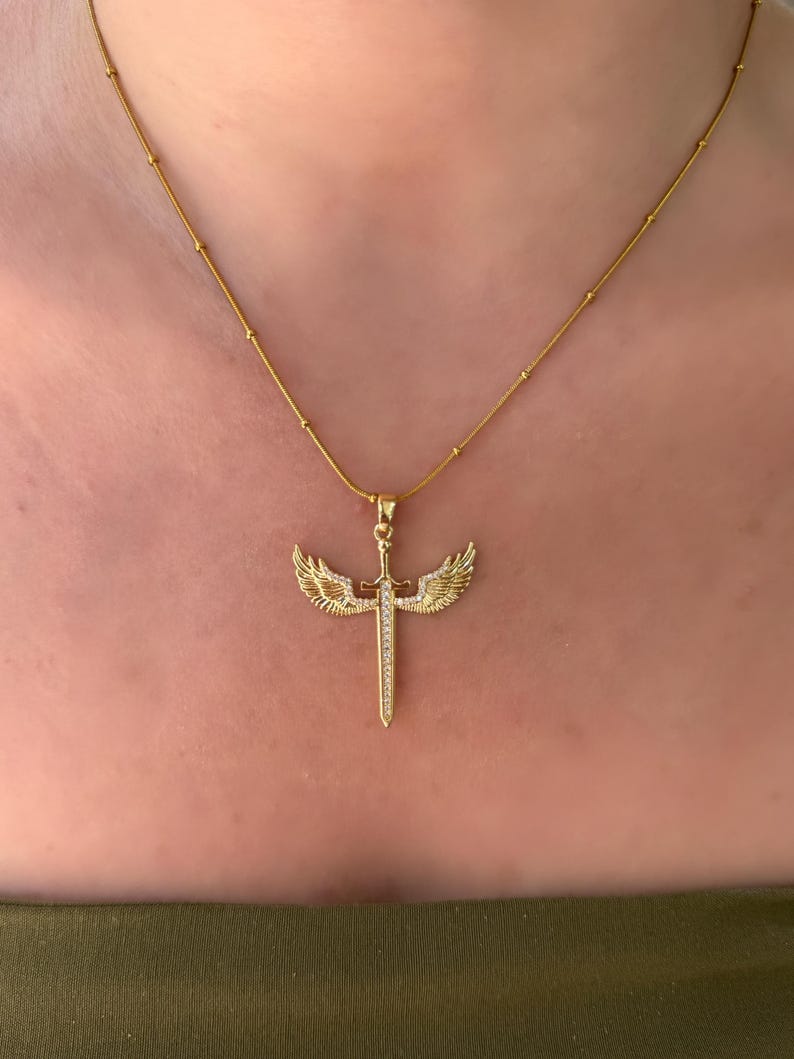 May include: A gold-colored necklace featuring a pendant with a sword design, adorned with small, sparkling stones. The sword is flanked by detailed wings, and the chain has small, evenly spaced beads. The necklace is displayed against a skin-toned background.