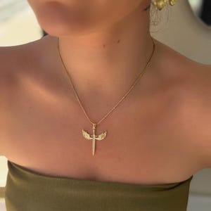 May include: A gold-colored necklace with a pendant shaped like a sword with wings. The pendant is encrusted with small, sparkling stones. The necklace is worn on a person wearing a green strapless top.