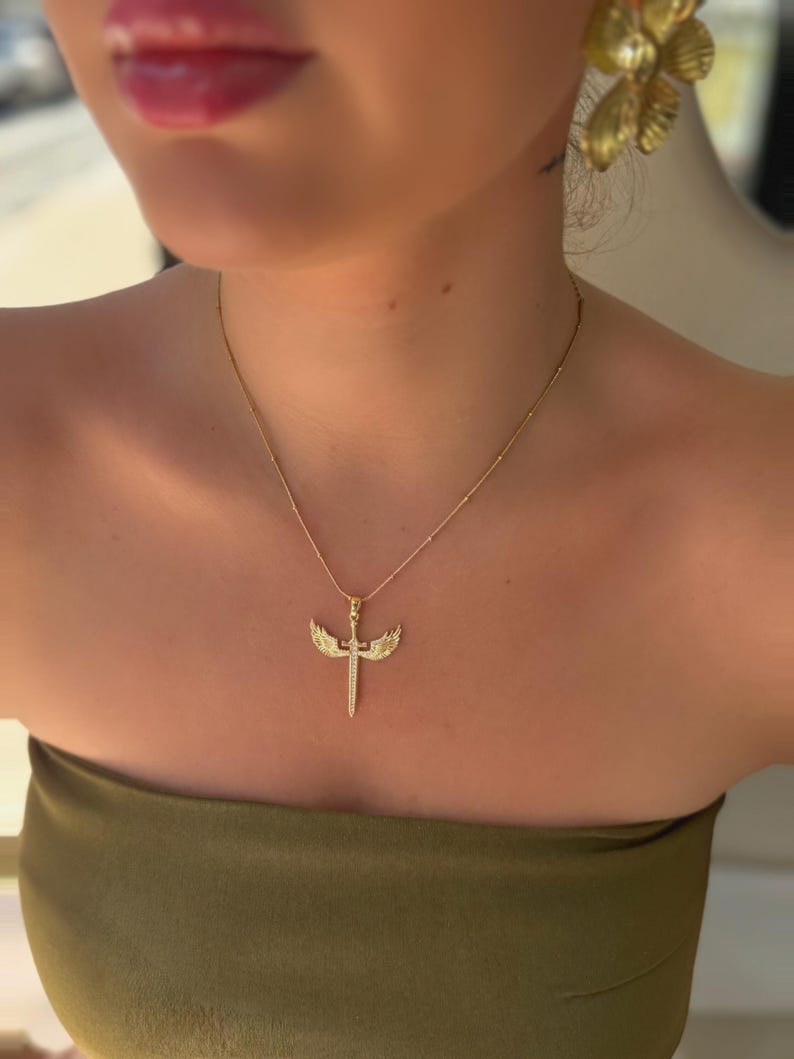 May include: A gold-colored necklace with a pendant shaped like a sword with wings. The pendant is encrusted with small, sparkling stones. The necklace is worn on a person wearing a green strapless top.