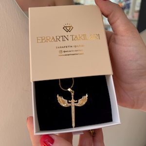 May include: A gold-colored necklace with a sword and wing pendant is displayed in a beige box. The box has gold lettering that reads "EBRAR'IN TAKILARI" and "ZARAFETIN ISILTISI". The necklace is on a black velvet background.