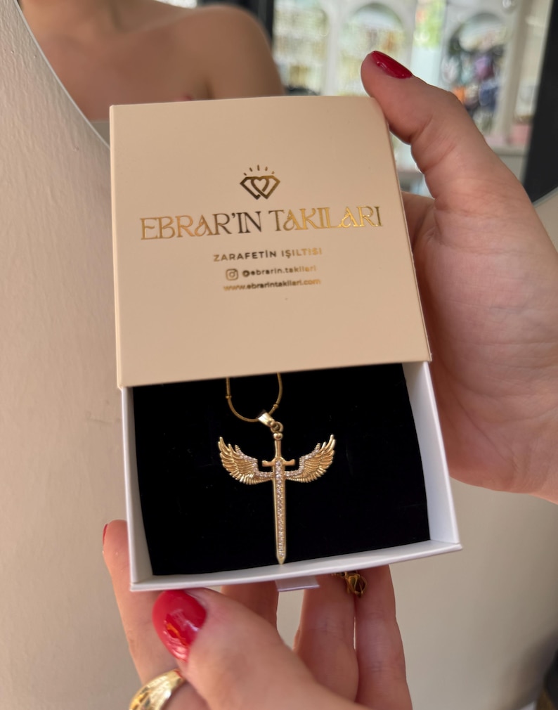 May include: A gold-colored necklace with a sword and wing pendant is displayed in a beige box. The box has gold lettering that reads "EBRAR'IN TAKILARI" and "ZARAFETIN ISILTISI". The necklace is on a black velvet background.