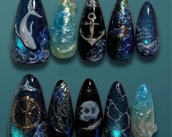 Puede incluir: Un conjunto de diez uñas artificiales largas en forma de almendra con temática náutica. Los diseños incluyen una ballena, un ancla, un timón y una calavera, todo en tonos azules, blancos y acentos iridiscentes. Las uñas tienen un acabado brillante.