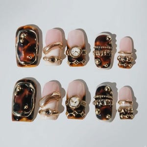 May include: A set of ten press-on nails featuring a mix of tortoiseshell patterns and solid light pink bases. The designs are embellished with gold-toned metal accents, including small spheres, textured bands, and a miniature clock face.