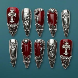 May include: A collection of ten silver-toned nail covers with intricate designs. Some feature a deep red polish, while others showcase detailed embellishments like crosses, hearts, and floral patterns. The nail covers are displayed against a teal background.