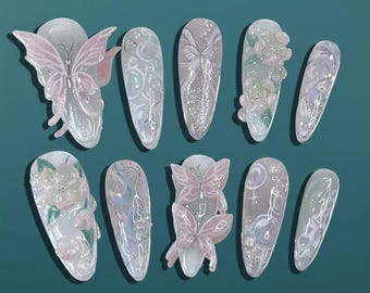 Puede incluir: Un conjunto de diez uñas artificiales con diversos diseños. Algunas uñas presentan adornos de mariposas y flores en 3D en tonos rosas y blancos. Otras uñas tienen patrones de purpurina y remolinos.