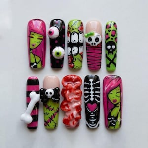 Puede incluir: Un juego de diez uñas postizas con diseños vibrantes que incluyen personajes de piel verde con cabello rosa, globos oculares en 3D, calaveras, huesos, una caja torácica con un corazón rosa, estampado de leopardo y patrones rojos texturizados.
