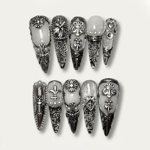 May include: A set of ten silver-toned, ornate nail designs. Each nail features intricate metalwork, including floral and cross motifs, with some nails showcasing glitter or pearl accents. The nails are arranged in two rows against a white background.