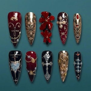 May include: A collection of ten long, pointed artificial nails. The nails feature various designs, including gold filigree, red floral accents, and pearl embellishments. Some nails are painted in deep red, gold, and black, with intricate patterns and textures.