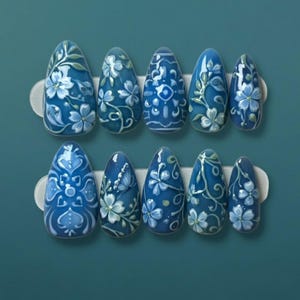 May include: A set of ten blue artificial nails with intricate floral designs. Each nail features a different pattern of white flowers and green vines, creating a cohesive yet unique aesthetic. The nails are arranged on a white holder against a teal background.