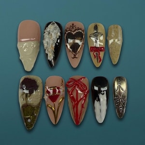 May include: A set of ten almond-shaped artificial nails with various designs. The designs include a skull, a feather, a heart, a sword, and a bow. The nails are painted in a variety of colors, including nude, black, gold, and red.