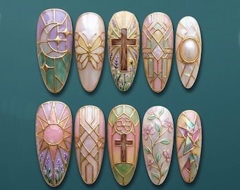 May include: A collection of ten oval-shaped nail art designs. Each nail features intricate designs with gold outlines, including crosses, floral patterns, and geometric shapes. The color palette includes pastel shades of pink, green, and white, with iridescent accents.