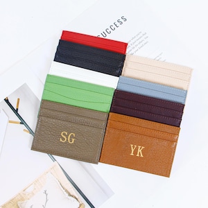 Puede incluir: Una colección de tarjeteros de cuero en varios colores, incluyendo rojo, negro, blanco, verde y marrón. Algunos tarjeteros tienen iniciales doradas. La palabra "SUCCESS" es visible en el fondo.