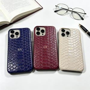 Pode incluir: Três capas de telemóvel com textura de pele de cobra nas cores azul marinho, bordô e creme. Cada capa tem iniciais douradas. Na imagem também estão óculos, uma caneta e um livro aberto. Um acessório elegante.