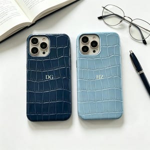 May include: Two phone cases with a crocodile skin texture. One is navy blue with the initials "DG", the other is light blue with the initials "HZ". The cases are next to a pair of glasses, a pen, and an open book.