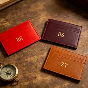 May include: Three personalized leather cardholders in red, burgundy, and brown, each with gold initials. A vintage compass sits nearby on a wooden surface. The cardholders have multiple card slots.
