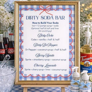 Op de afbeelding: Een ingelijst bord met een blauw-wit geruit patroon en een rode geschulpte rand. Het bord toont "DIRTY SODA BAR" en frisdrankrecepten. Frisdrankflessen, ijs en gearomatiseerde siropen zijn ook zichtbaar.