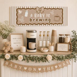 May include: A baby shower coffee station with a Keurig coffee maker, mugs, and syrup bottles. A banner reads "Baby on the Way." A sign says "A Baby is Brewing." The decor includes flowers, a teddy bear, and a candle.