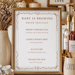 Può includere: Un cartello incorniciato con il testo "BABY IS BREWING DRINK SPECIALS". Il menu elenca "Mommy's Morning Fuel", "Daddy's Double Shot", "Ice Ice Baby" e "Snuggle Me Chai". La cornice è dorata e lo sfondo è bianco a righe.
