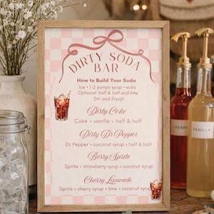 May include: A framed sign with the text "DIRTY SODA BAR" in a pink and white checkered design. The sign includes drink recipes like "Dirty Coke" and "Berry Sprite." Two syrup bottles are visible, labeled "CHERRY" and "VANILLA."