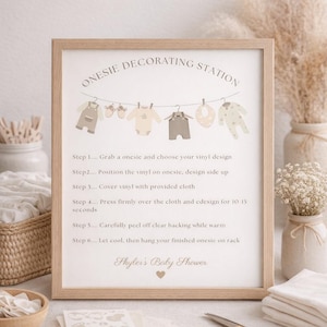 Op de afbeelding: Een ingelijst bord met de tekst "Onesie Decorating Station" met instructies en illustraties van babykleding. Een schaar, vinylontwerpen en opgevouwen doeken zijn ook zichtbaar.