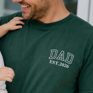 May include: A dark forest green short-sleeved t-shirt featuring white embroidered text on the left chest area. The text displays the word DAD in a bold collegiate font, with the smaller text EST. 2026 positioned directly underneath it.