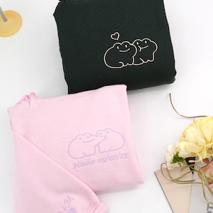 Peut inclure: Deux sweat-shirts, un vert foncé et un rose, sont présentés. Le sweat-shirt vert foncé présente un motif brodé blanc de deux grenouilles et un cœur. Le sweat-shirt rose a un motif de grenouille assorti, le nom "Minho 05/07/23", et les initiales "A+M" brodées sur la manche.