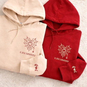 May include: Two hooded sweatshirts, one in beige and one in red, are shown. Each has a heart-shaped spiderweb design and the Roman numeral date II.XIV.MMXXV. The beige sweatshirt has the letters "Q&L" embroidered on the cuff.