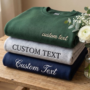 May include: A stack of three folded sweatshirts in green, gray, and navy blue. Each sweatshirt has the words "custom text" embroidered on the front in a contrasting color. The sweatshirts are stacked on a wooden surface.