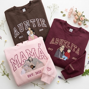 Pode incluir: Três camisolas de gola redonda em castanho, bordô e rosa. Cada camisola apresenta texto bordado: "AUNTIE", "ABUELITA" e "MAMÁ", com ilustrações familiares e o ano de fundação. Acentos florais circundam as camisolas.