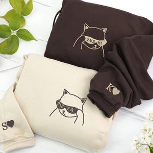 May include: Two sweatshirts, one cream and one brown, with cat graphics. The cream sweatshirt has "PIZZA" on the cat's blindfold and an "S" with a heart on the sleeve. The brown sweatshirt has "SUS HI" on the cat's blindfold and a "K" with a heart on the sleeve.