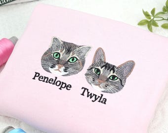 May include: A light pink square pillow featuring embroidered cat faces with green eyes. The names "Penelope" and "Twyla" are embroidered below the cat faces. The pillow is on a white surface.