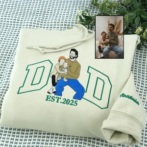 May include: Cream-colored hoodie with a green embroidered design. The design features the word "DAD" in a collegiate font, a family illustration, and the text "EST.2025". The sleeve has the name "Anastasia" embroidered in green. A small framed photo is in the upper right corner.
