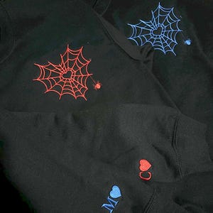 Black couple hoodies with custom embroidered spider web and heart design on the chest in red and blue, with matching initials and heart detail on the sleeve. Unique Halloween-themed sweatshirts, personalized embroidery gift for spooky season, romantic gothic couples outfit, cozy and stylish for fall or October matching look.