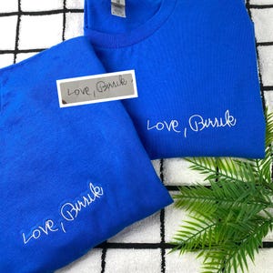 May include: Two royal blue t-shirts with the embroidered text "Love, Buruk" in white. One shirt has the text on the front, and the other has the text on the sleeve. A small white label with the same text is also visible.