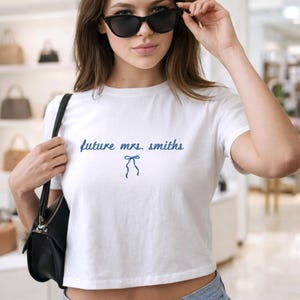 May include: White crop top featuring the text "future mrs. smiths" in blue script, and a blue bow graphic. The top is a casual style.