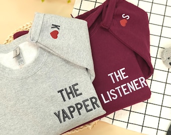 Puede incluir: Se muestran dos sudaderas, una gris y otra burdeos. La sudadera gris tiene impreso "THE YAPPER", con un corazón rojo y la letra "K" bordada en la capucha. La sudadera burdeos tiene impreso "THE LISTENER", con un corazón rojo y la letra "S" bordada en la capucha.