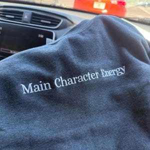 Heather Dark Navy sweatshirt displayed inside a car, featuring white embroidered text reading “Main Character Energy.” The phrase is stitched in a clean serif font across the chest, with subtle raised embroidery that contrasts softly against the heather gray fabric.