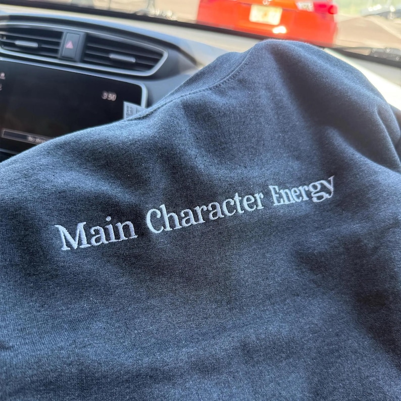 Heather Dark Navy sweatshirt displayed inside a car, featuring white embroidered text reading “Main Character Energy.” The phrase is stitched in a clean serif font across the chest, with subtle raised embroidery that contrasts softly against the heather gray fabric.