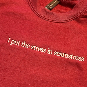 Red Gildan crewneck sweatshirt with white embroidered text saying “I put the stress in seamstress.” The lettering is stitched in a simple lowercase font across the front chest, creating a playful sewing-themed quote with clean embroidery and visible thread texture.