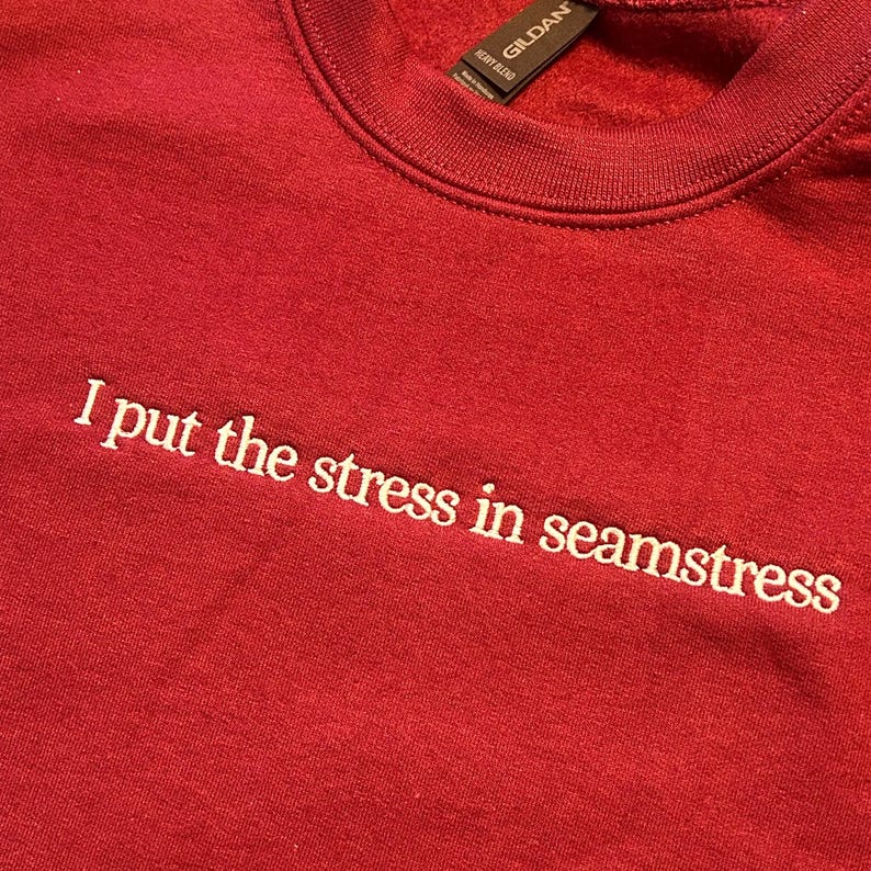 Red Gildan crewneck sweatshirt with white embroidered text saying “I put the stress in seamstress.” The lettering is stitched in a simple lowercase font across the front chest, creating a playful sewing-themed quote with clean embroidery and visible thread texture.