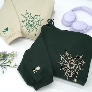 Sand and Forest green hoodies styled on a white surface with accessories, featuring embroidered spider web heart design on the chest and a small spider detail at the bottom. Sleeve embroidery includes custom initial letters with a heart symbol. Modern minimalist embroidery, personalized couple hoodies, unique Valentine or anniversary gift.