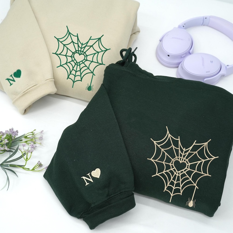 Sand and Forest green hoodies styled on a white surface with accessories, featuring embroidered spider web heart design on the chest and a small spider detail at the bottom. Sleeve embroidery includes custom initial letters with a heart symbol. Modern minimalist embroidery, personalized couple hoodies, unique Valentine or anniversary gift.