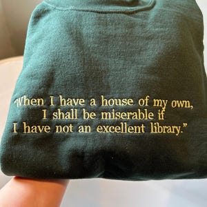 Forest green embroidered sweatshirt featuring a gold thread quote that reads “When I have a house of my own, I shall be miserable if I have not an excellent library.” The text is stitched in a classic serif font, evenly spaced across the chest, giving a literary and vintage-inspired aesthetic.
