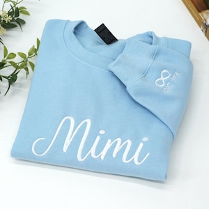 Light blue embroidered sweatshirt folded neatly on a white surface, featuring large white cursive embroidery spelling the name “Mimi” across the front chest. The script font is smooth and elegant, with thick satin stitching that stands out clearly on the soft pastel blue fabric. Minimal styling highlights the personalized name embroidery.