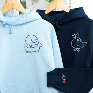 May include: Two hooded sweatshirts, one light blue and one navy. The light blue sweatshirt features a cartoon penguin holding a knife. The navy sweatshirt has a white duck graphic. The navy sweatshirt sleeve has the initials "AB" and a red heart.